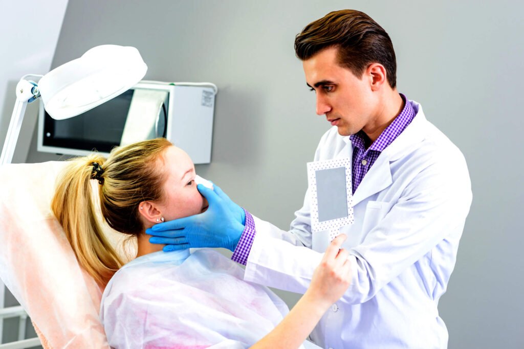 A dentist holds a mirror and examines a patient's mouth while covering it with his gloved hand.