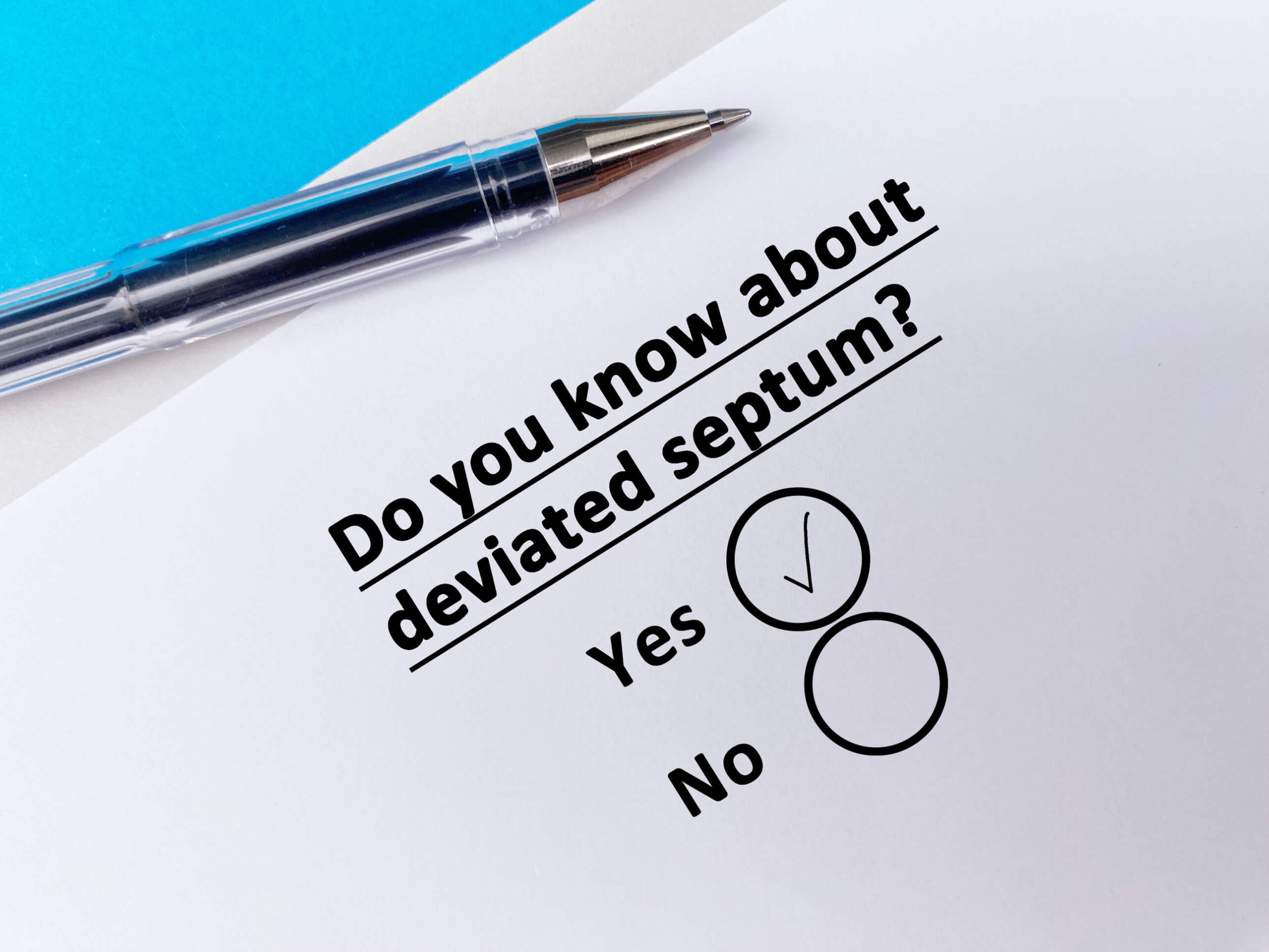 A questionnaire with the question "Do you know about deviated septum?" and two options: "Yes" with a check mark and "No" in an empty circle, lies beside a pen. There's also a note asking, "Is facelift surgery covered by insurance?